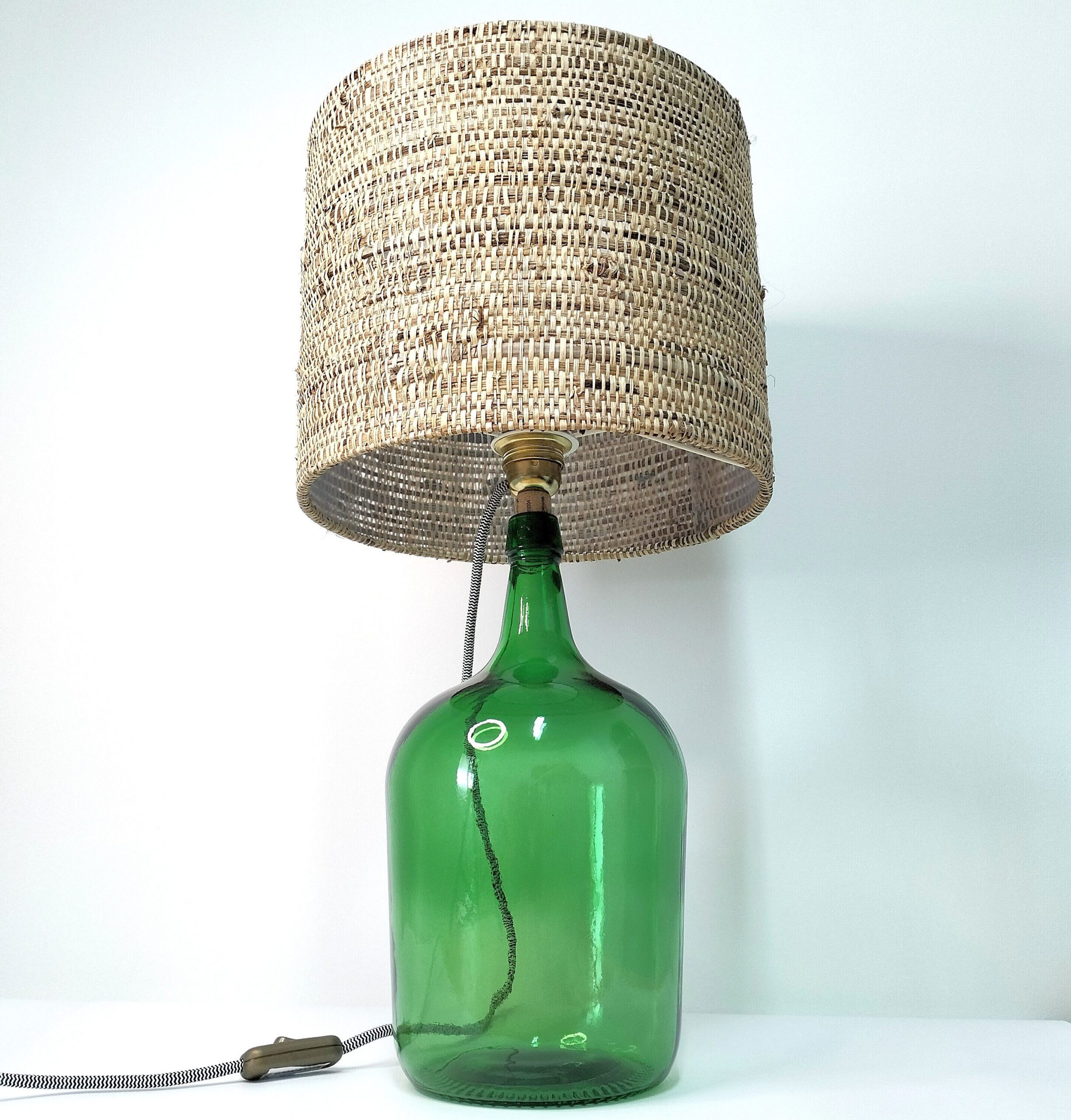Este candeeiro de mesa é a combinação perfeita de iluminação funcional e beleza decorativa. Candeeiro de mesa, base de vidro, abajur palhinha. Está sobre um fundo branco.