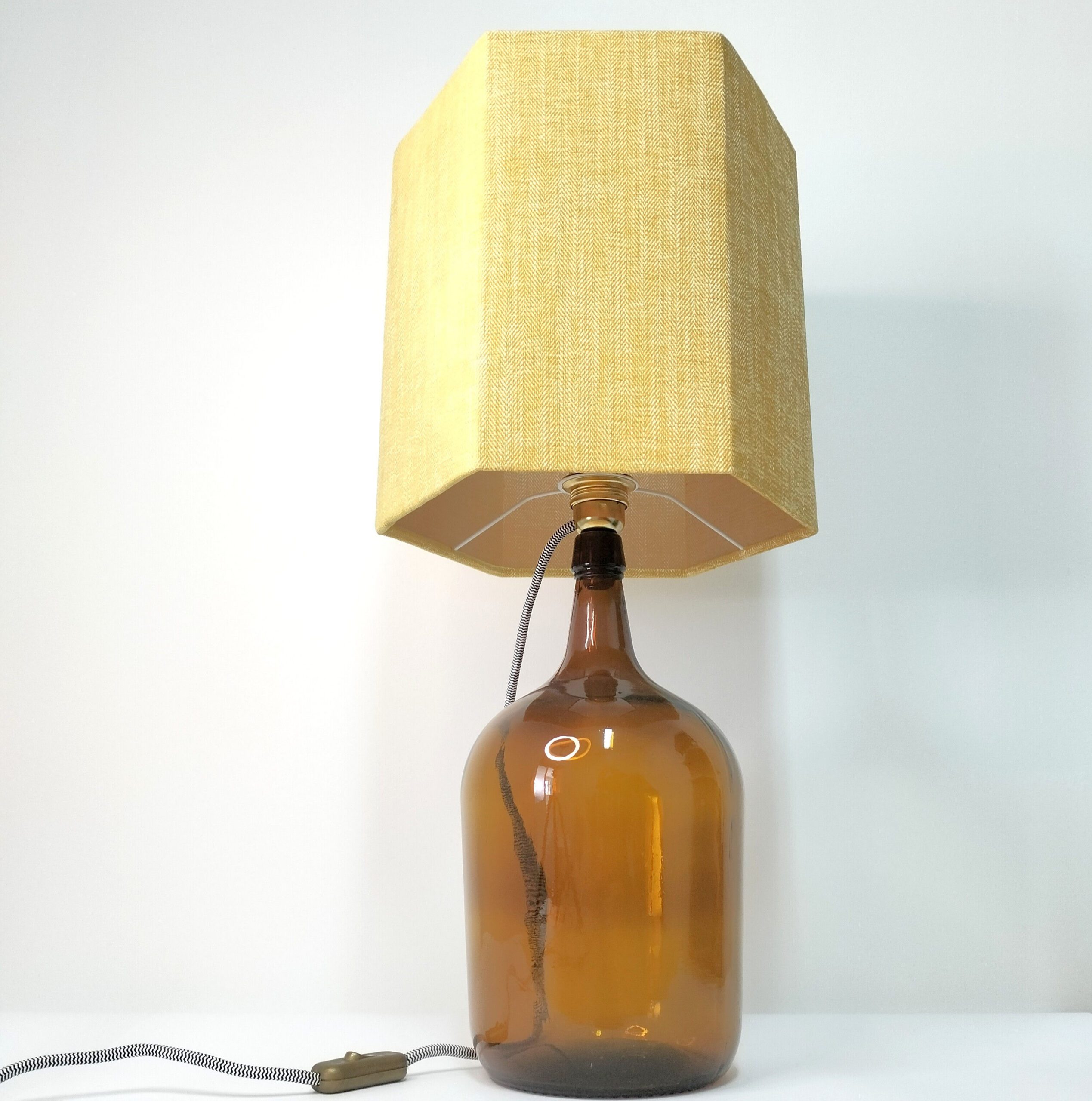 Este candeeiro de mesa une praticidade e estilo numa peça única. O seu design moderno é perfeito para criar um ambiente aconchegante enquanto complementa sua decoração com personalidade. A base é um garrafão de vidro castanho e o abajur amarelo. Está sobre um fundo branco.