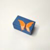 Caixa artesanal azul com borboleta laranja, tamanho mini. Está exposta fechada sobre um fundo branco.