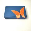 Caixa artesanal azul com borboleta laranja. Está sobre um fundo branco.