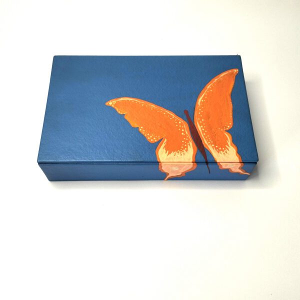 Caixa artesanal azul com borboleta laranja. Está sobre um fundo branco.