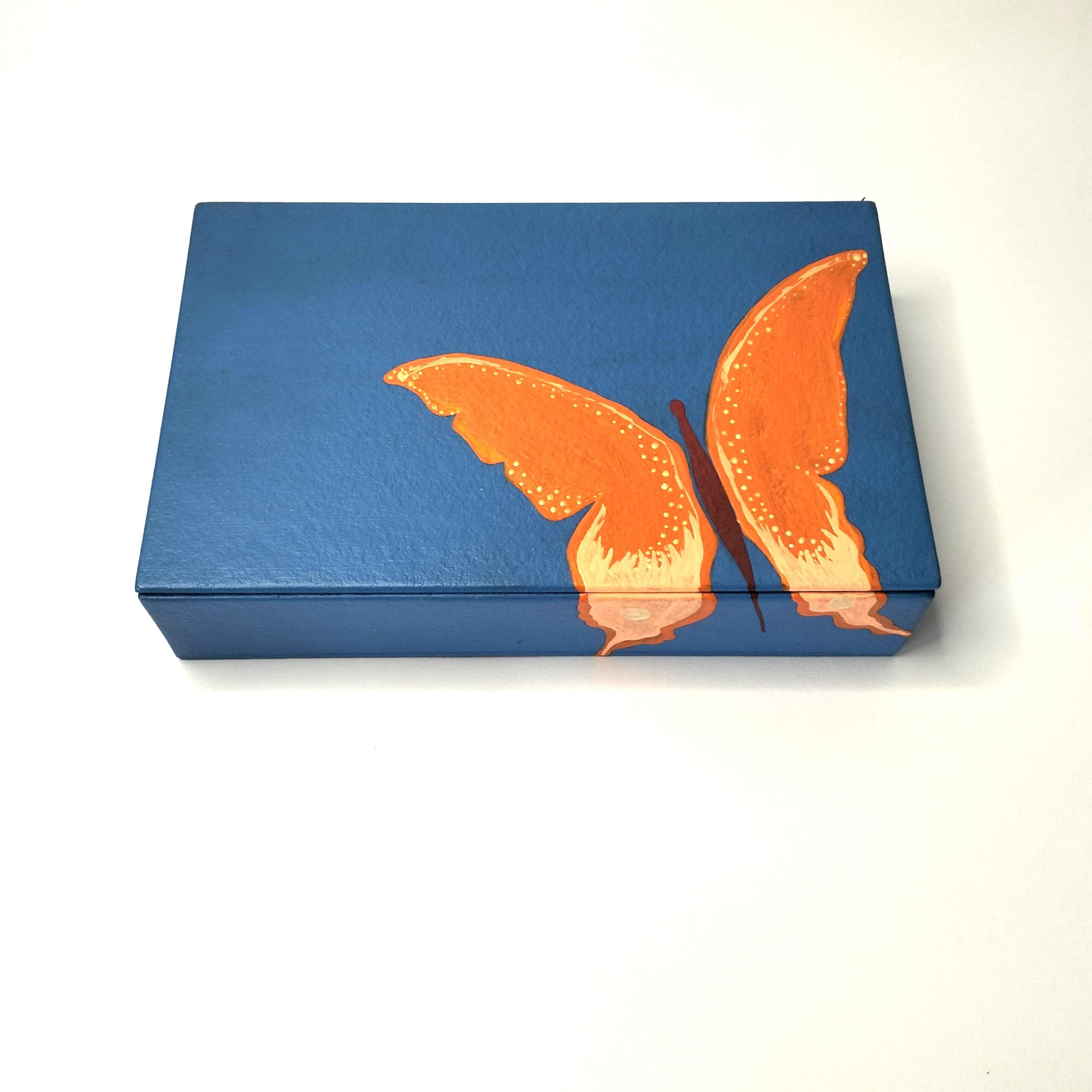 Caixa artesanal azul com borboleta laranja. Está sobre um fundo branco.