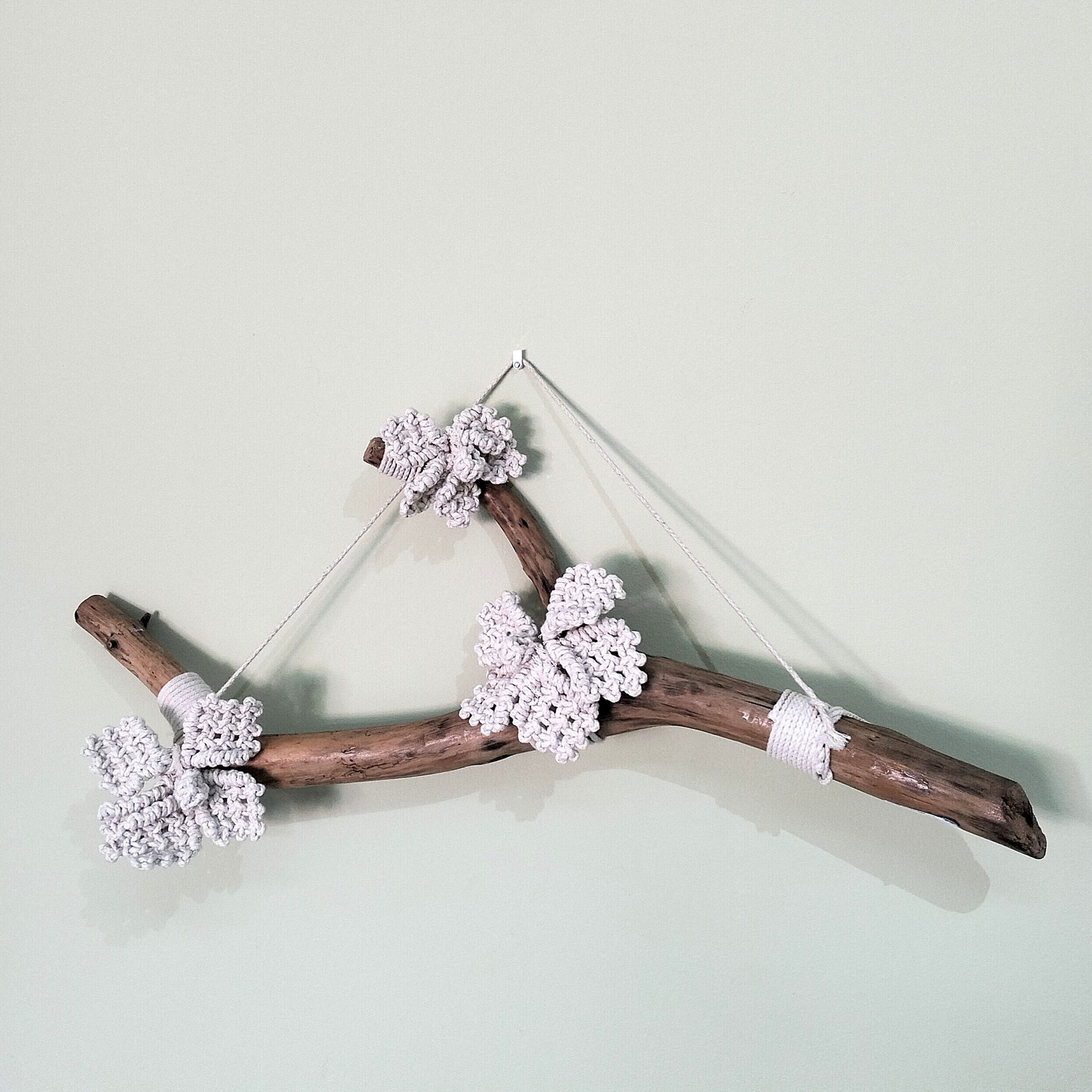 Macramé artesanal em tronco para pendurar. Esta peça decorativa combina a rusticidade do tronco com a delicadeza do macramé. Está exposto sobre um fundo branco.