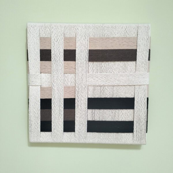 Quadro artesanal com fios de algodão em tons de creme, castanho e preto. Está exposto numa parede verde claro.