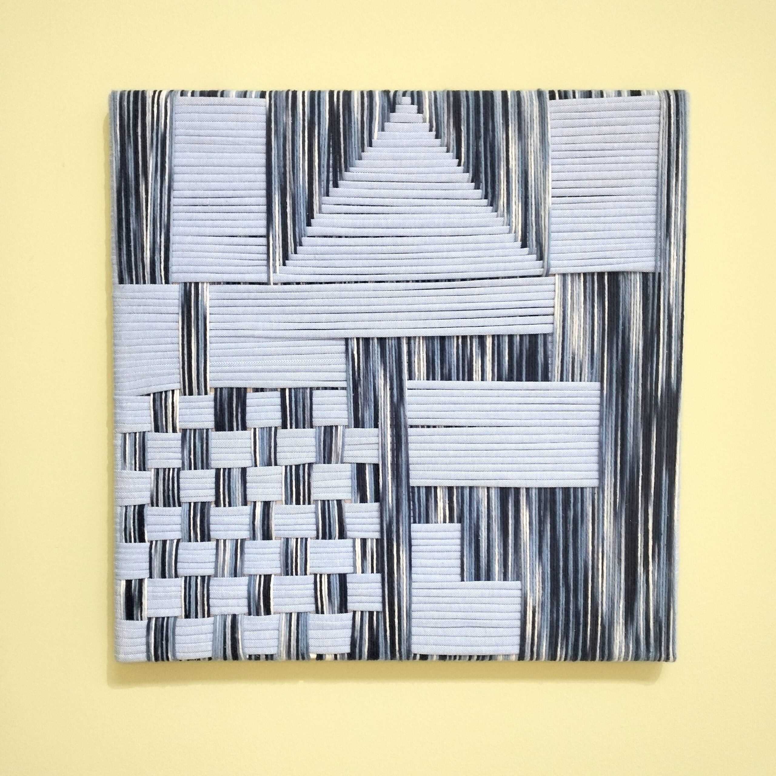 Quadro artesanal com fios de algodão azul. Está exposto numa parede amarela.