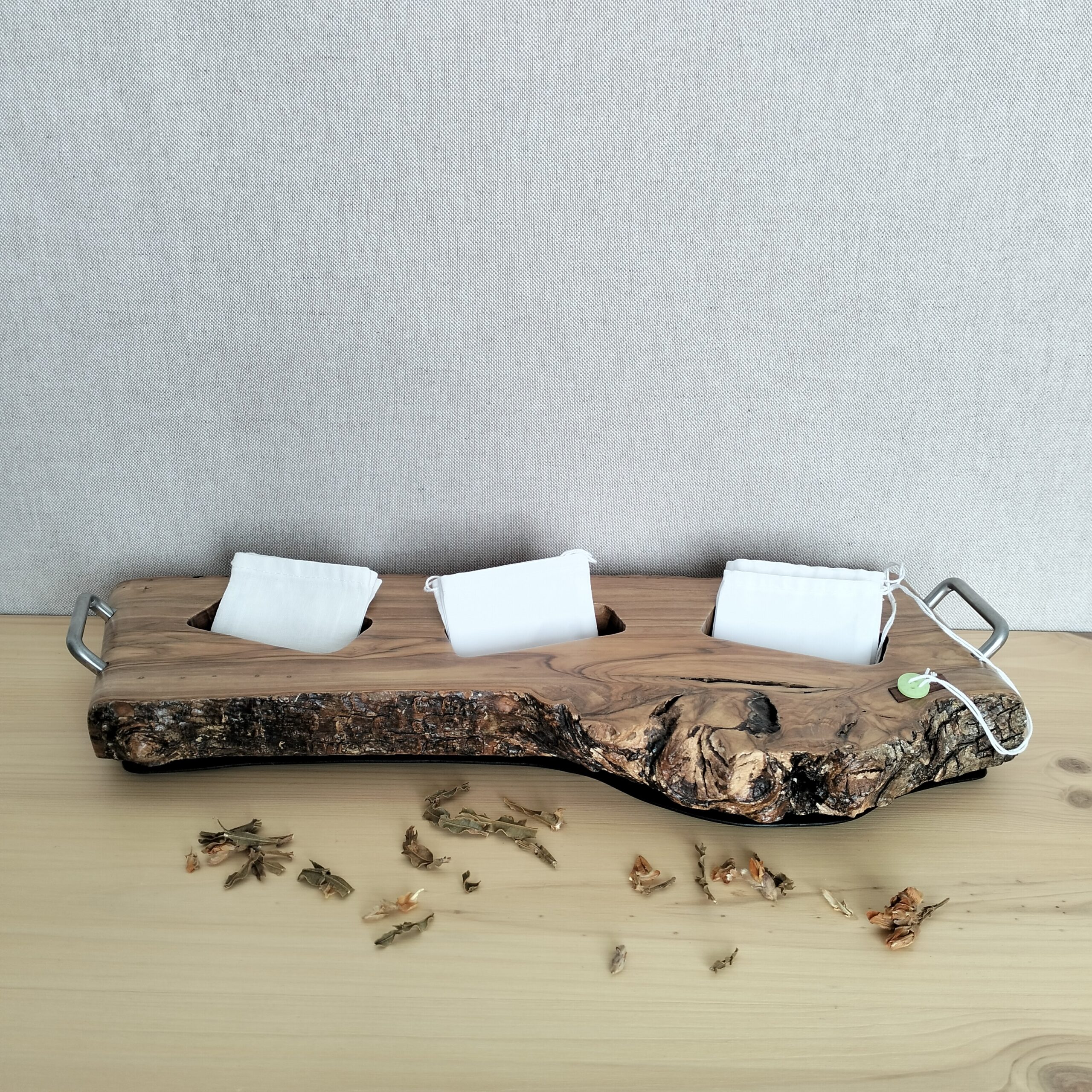 Suporte artesanal para saquetas de chá em madeira de oliveira e pagas em inox. Está exposto numa prateleira de madeira clara, com saquetes de chá algodão e chá a decorar. Tudo com fundo de cor neutra.
