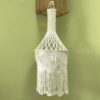 Candeeiro de teto artesanal em macramé no tom natural do algodão. Está pendurado sobre um fundo verde.