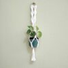 Suporte artesanal para vaso em macramé branco. Está suspenso com um vaso azul e planta verde dentro.