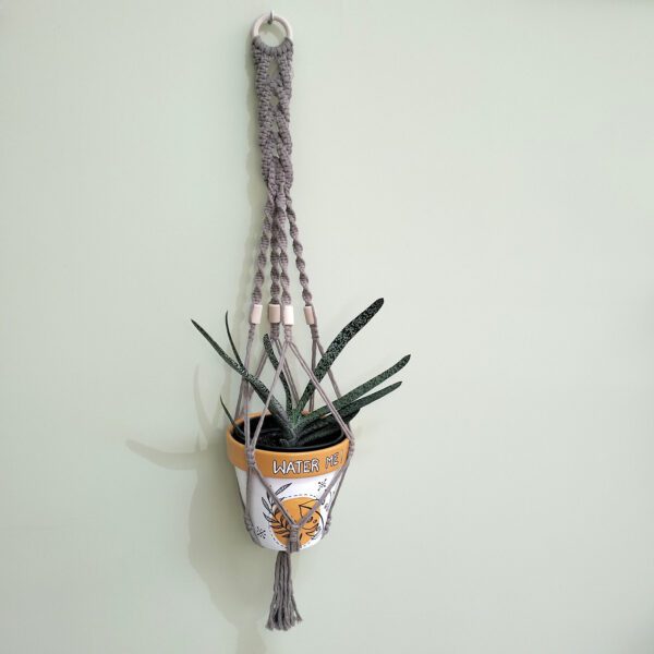 Suporte artesanal para vaso em macramé bege com contas e argola de madeira. Está suspenso numa parede com um vaso com uma suculenta dentro.