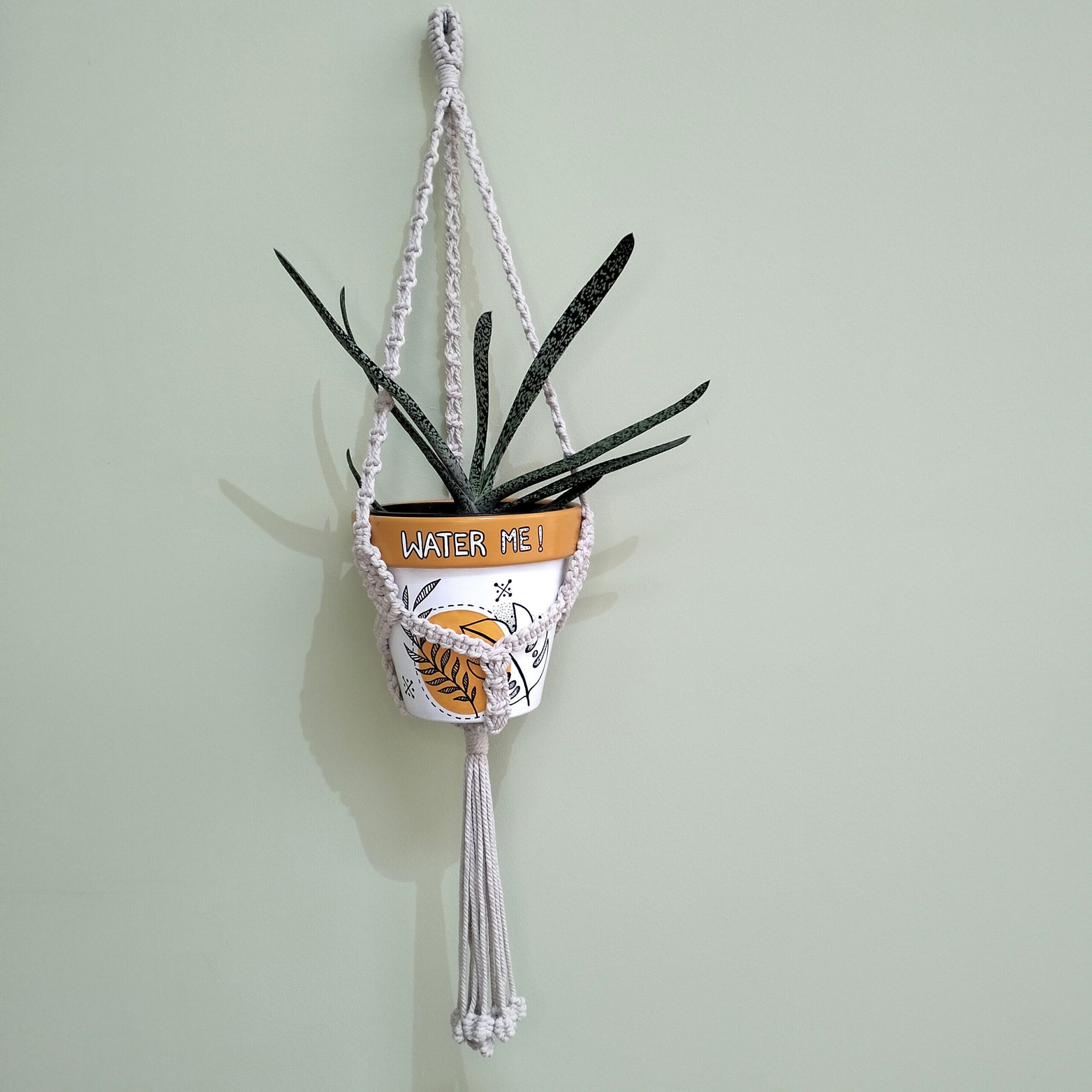 Suporte para vaso artesanal em macramé natural com bolas nas pontas, também elas feitas em macramé. O suporte está pendurado junto a uma parede com um vaso de teraacota pintado à mão em tons de laranja e uma suculenta dentro.