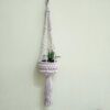 Suporte para vasos artesanal em macramé com pompons. Está suspenso junto a uma parede com um vaso com uma suculenta dentro.