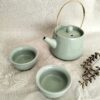 Bule e duas chávenas de chá verde-água. Estão com um fundo de tecido bege e um raminho seco a decorar o ambiente.