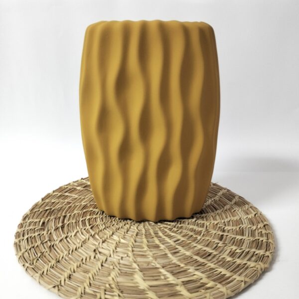Jarra artesanal de cerâmica amarelo mostarda, com efeito de ondas. Está apresentada num fundo branco sob uma base redonda de folha de palma.