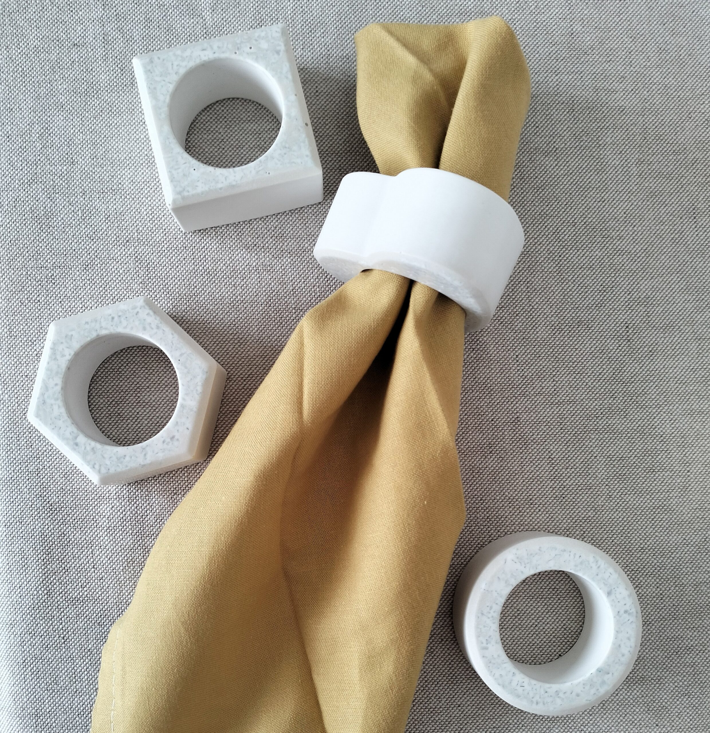 Quatro argolas para guardanapos (coração, redonda, quadrado e hexagonal) em resina ecológica com granulado de vidro reciclado verde. Conjunto artesanal, está sobre um fundo bege e uma das argolas envolve um guardanapo de pano amarelo.