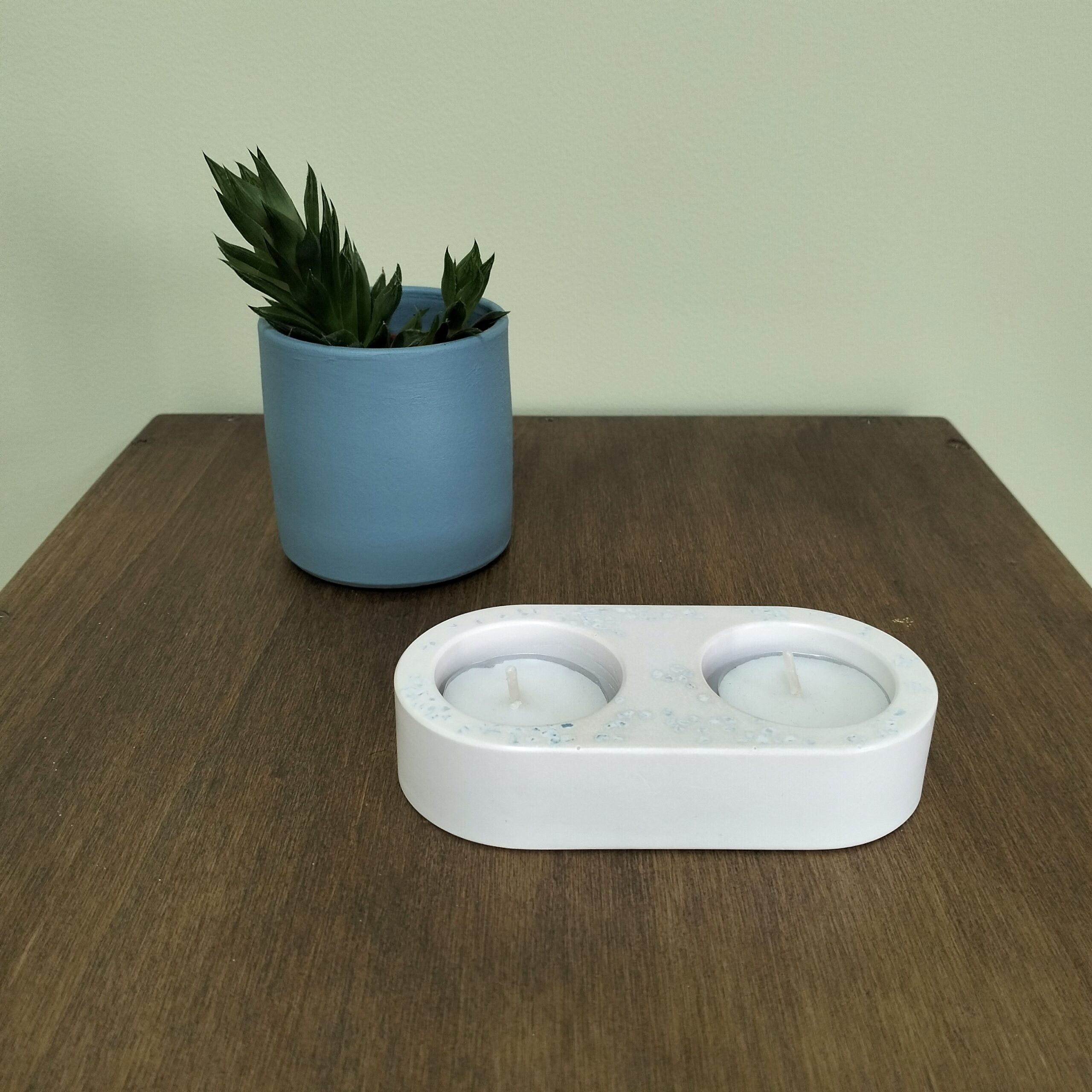 Castiçal para 2 vela tealight em resina ecológica em tons de areia. Está sobre madeira com uma vaso de cerâmica azul com suculenta atrás.