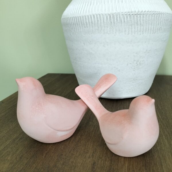 Conjunto de dois pássaros de tamanhos diferentes, em resina ecológica na cor coral. Estão expostos de costas um para o outro com os rabos a tocarem-se. Estão sobre madeira com uma jarra de cerâmica no fundo.