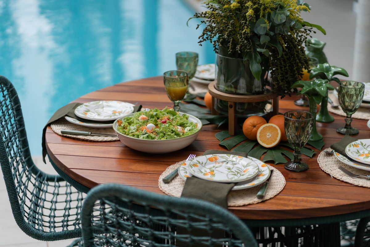 Ambiente de refeição ao ar livre. Mesa redonda de madeira decorada em tons de verde e laranja, com flores e com água da piscina como fundo.