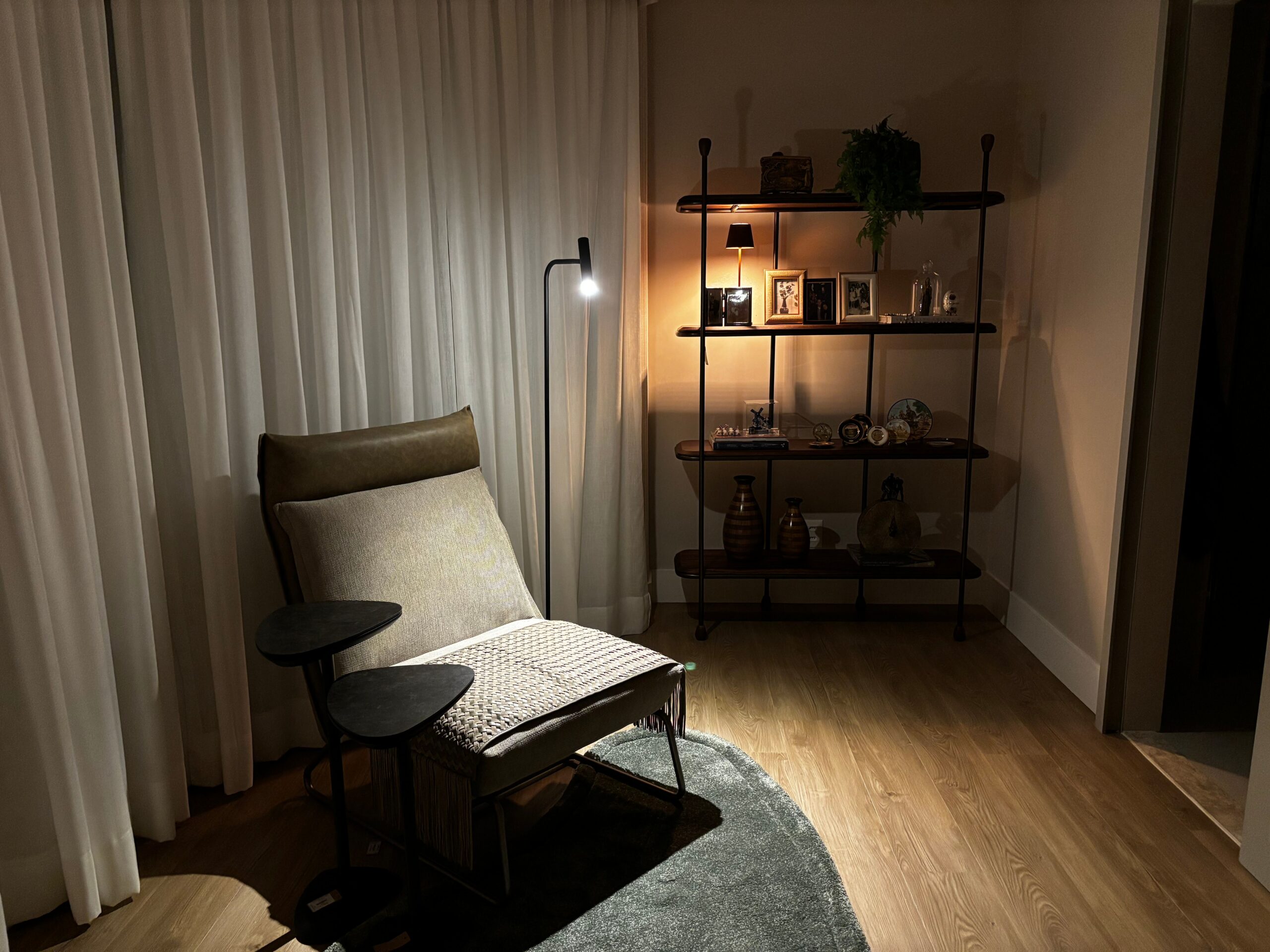 Canto de leitura fotografado de noite, há um candeeiro de pé com luz branca funcional para a leitura e atrás uma estante com uma luz amarela, acolhedora e decorativa.