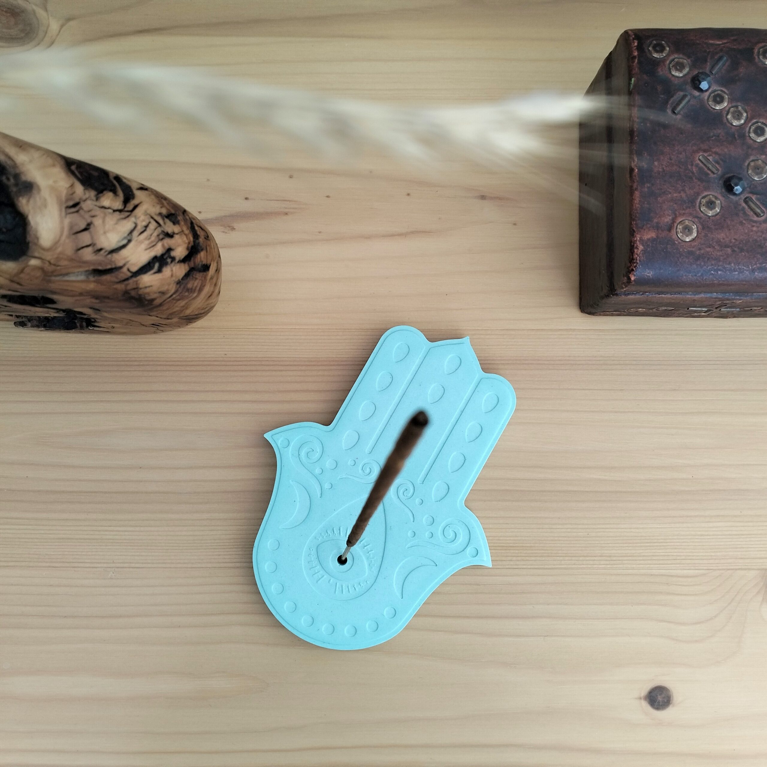 Suporte para incenso em forma de Mão de Fátima ou Hamsá, feito em resina ecológica num tom suave de verde água. Está sobre um móvel com uma caixa e uma jarra de madeira dos lados.