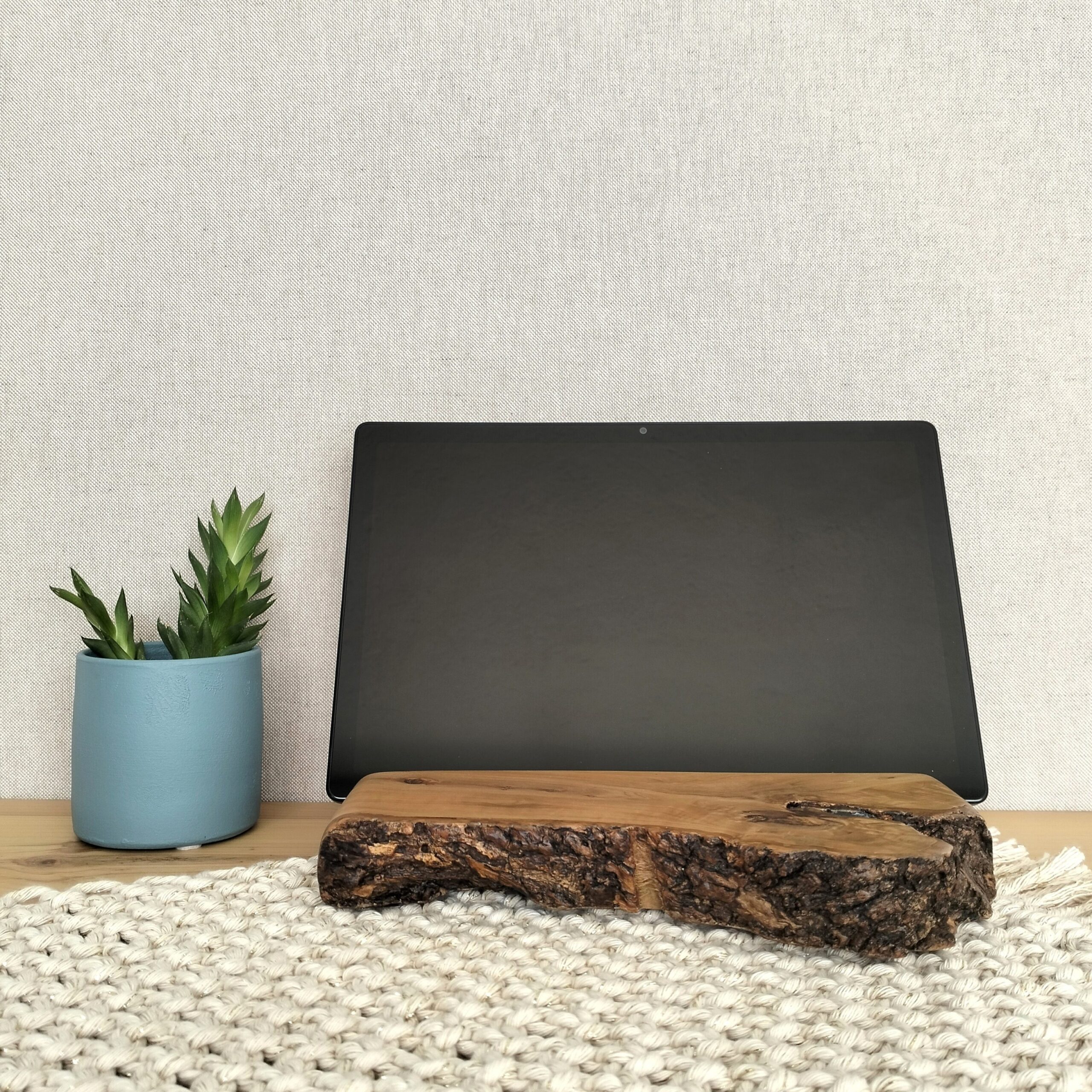 Tablet colocado num suporte artesanal em madeira de sobreiro adequado para o efeito. Está sobre uma peça de macramé e um pequena planta ao lado.